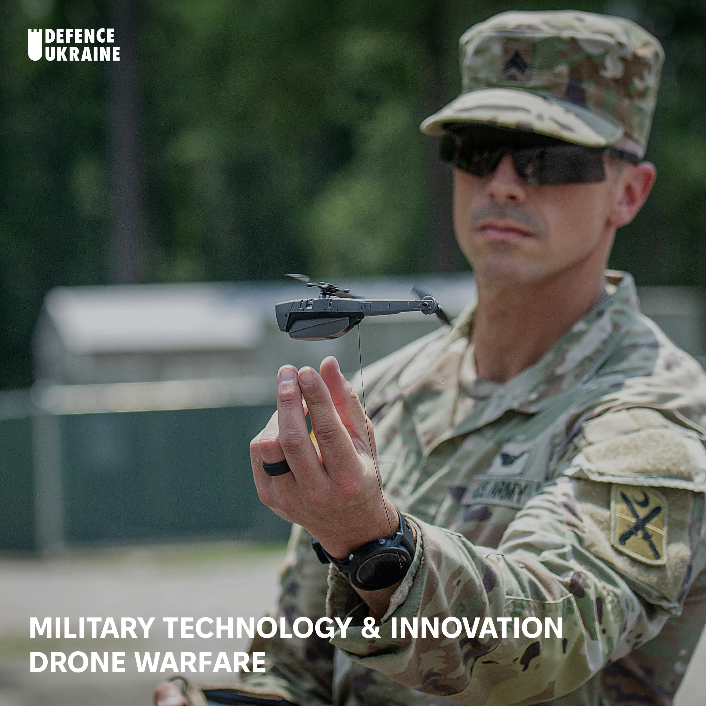 A US Army soldier holds a Teledyne FLIR Black Hornet nano-drone between his fingers during a training exercise, showing the 33-gram helicopter actual size against a human hand.